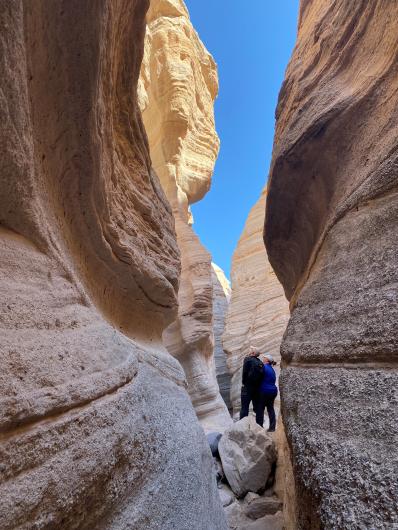 Two individuals navigate a narrow canyon in the desert, flanked by steep rock walls and a clear blue sky above.