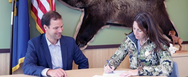 BLM AK State Director Kevin Pendergast and DOI Deputy Secretary Kate MacGregor sit at a table in front of the Alaska and American flags and a hung bear pelt signing a piece of paper.