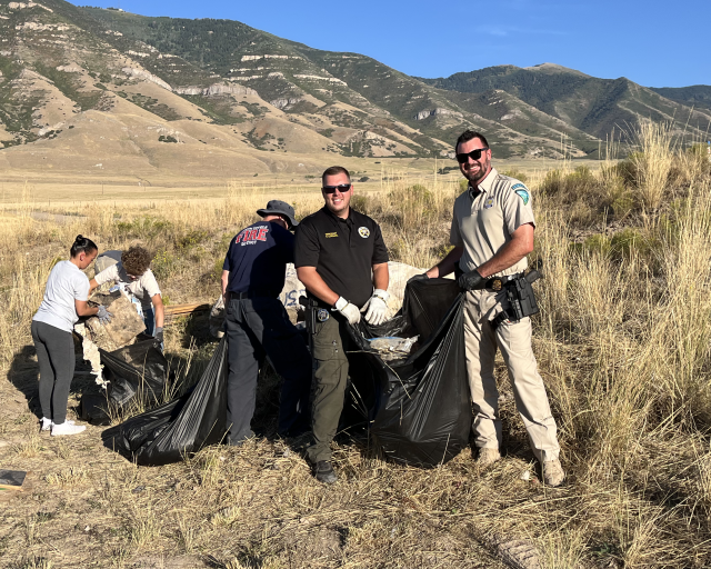 A BLM Law Enforcement Ranger and a Tooele County Sherrif's Department officer place trash on a trash bag with other persons in the background 
