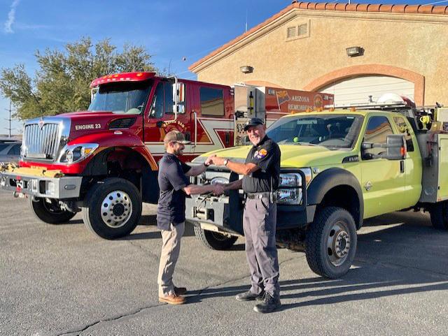 Two men exchange keys in front of fire engines.