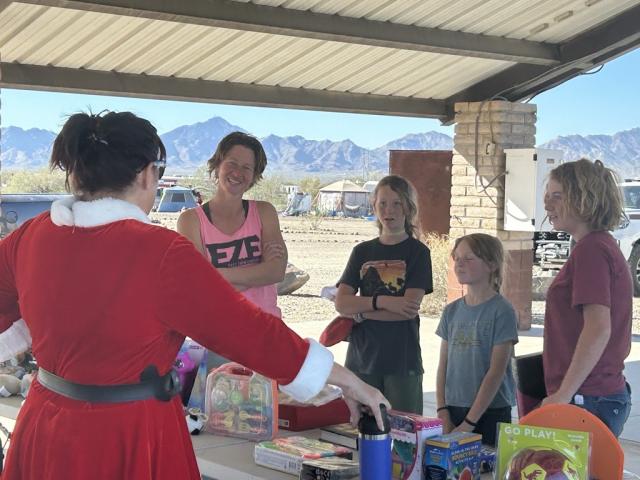 Mrs. Claus with her back to the camera visits with a family at the gift table.
