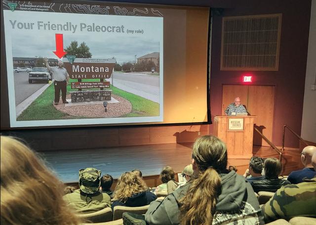 Audience watching a speaker presenting a slide titled 'Your Friendly Paleocrat' showing a man by a Montana State Office sign with a red arrow pointing at him.