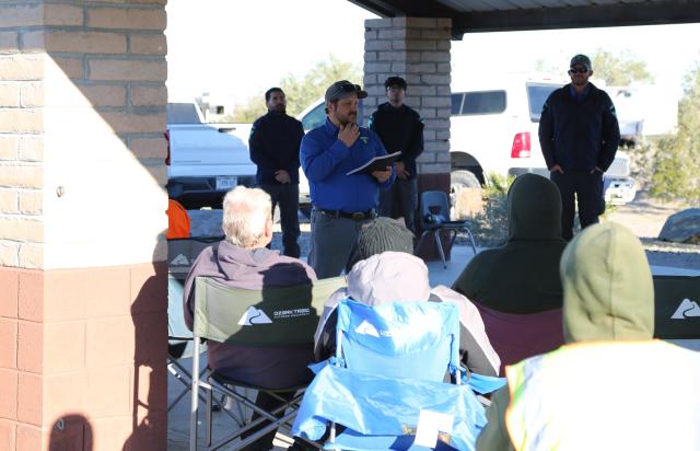 A blue clad park ranger with a grey BLM hat addresses a group of volunteers.