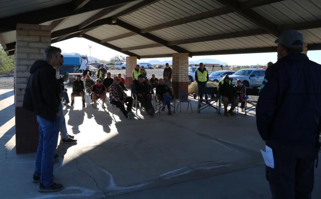 A group of approximately twenty volunteers gather under a shade structure for volunteer training. Most are sitting but some standing.