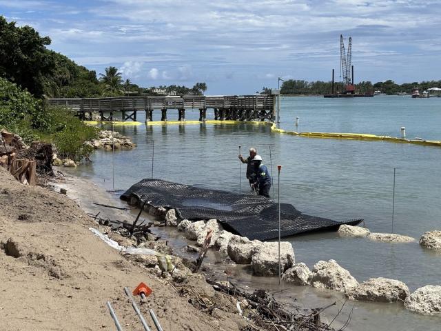 Two workers on a beach are adjusting a black net in shallow water, with a wooden pier and construction equipment in the background.