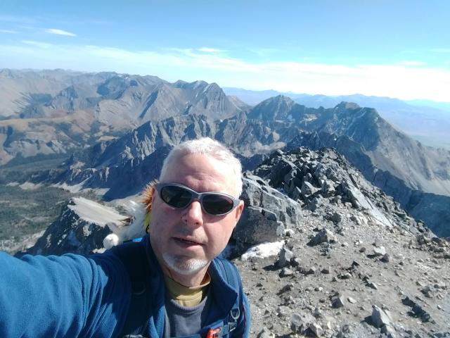 A person stands on a mountain peak, surrounded by rugged terrain and distant peaks under a clear blue sky.