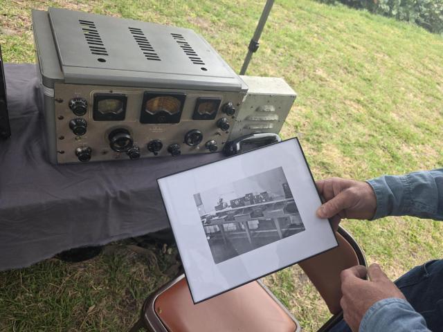 Person holding a framed black-and-white photo next to a vintage radio device on a table outdoors with grass in the background.