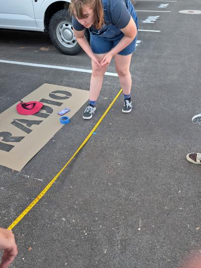 A young woman uses a tape measure in a black parking lot to paint parking lines