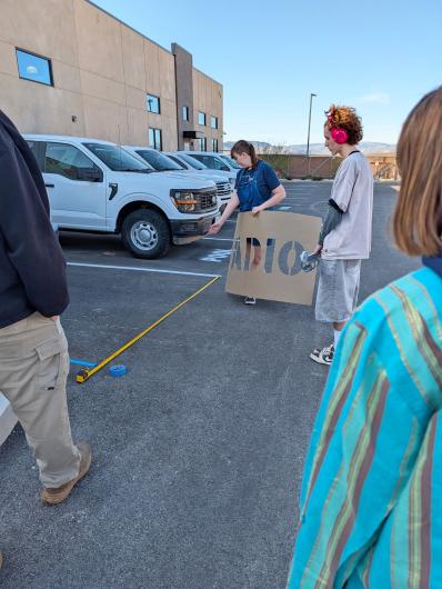 Youth hold a giant brown cardboard stencil to paint white lines on a black parking lot