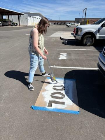 A young woman lays a brown cardboard stencil on a black parking lot to prepare to paint letters in white