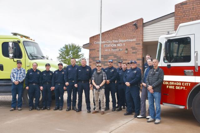 A large group of firefighters stand in front of a red and a green fire truck