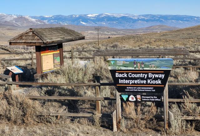 A sign with the logos for BLM’s National Conservation Lands and Back Country Byways programs and the Lewis and Clark Historic National Trail sits next to a shaded kiosk with site and safety information, with additional interpretive signs nearby. A fence made of weathered wood surrounds the kiosk, with sagebrush growing in the foreground and middle distance. A mountain range topped with patches of snow appears in the far distance. 