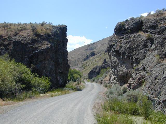 A smooth gravel road wide enough to accommodate two vehicles traveling in opposite directions provides a gently curving route between two cliff faces in the middle distance. Shrubs, grasses, and other vegetation grow on both sides of the road, which skirts a steep hillside in the far distance. Overhead is a bright blue sky with a few large white clouds. 