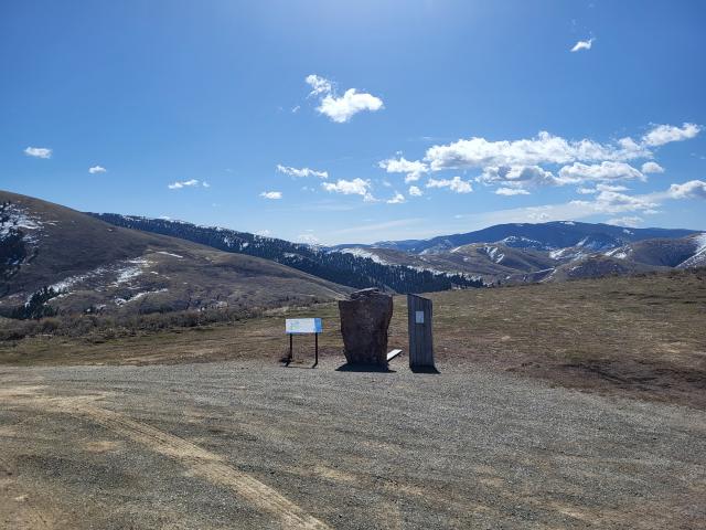 A gravel pullout provides access to an interpretive sign and two other historical markers on the side of the byway. In the middle and far distance mountains with patches of snow appear under a bright blue sky with small, wispy clouds scattered across it. 