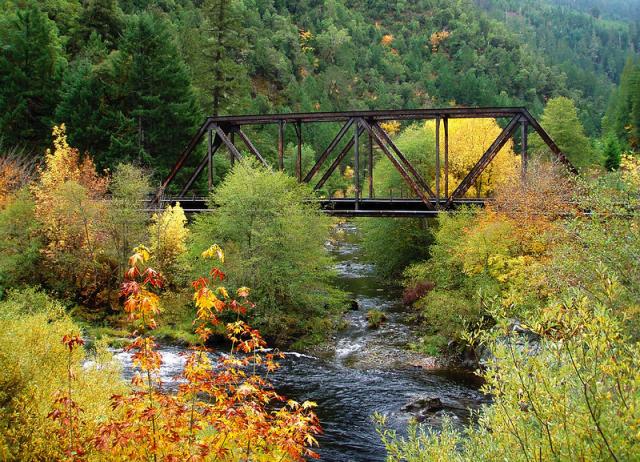 A metal truss bridge crosses over a shallow, rocky section of the Creek, whose dark waters and riffles are bordered by the greens, yellows, oranges, and reds of autumn foliage.  