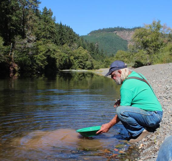  man wearing jeans, a green T-shirt, suspenders, and a blue baseball cap crouches on a riverbank while holding over the water a wide, flat pan that matches the color of his shirt. Sediment suspended in the water reveals that the pan is being used to sift gold particulate from bottom silt. 