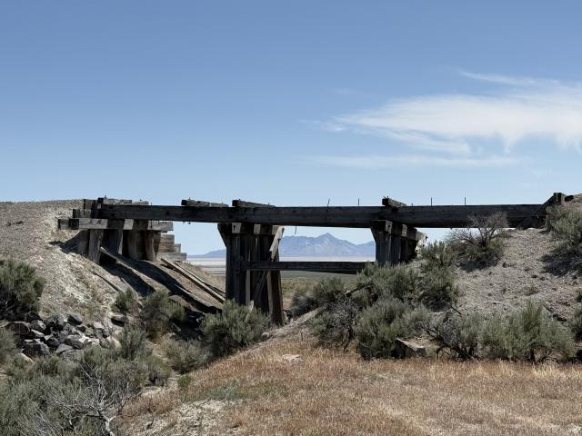 Wooden trestles support the remnants of the deck of a railroad bridge that crosses over a gulch. Short brown grass and small green shrubs grow in the foreground and middle distance; beyond the bridge, in the far distance, part of a mountain range appears beneath a nearly cloudless blue sky. 