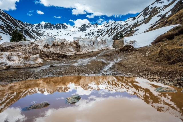 A dirt road with rocks, icy patches, and an uneven surface passes between a snow-rimmed, water-filled declivity in the foreground and snowy embankments in the middle distance. Farther away, brownish-red mountains covered in patches of snow have a color suggestive of cinnamon. A large, pillowy cloud hanging over the mountains is reflected in the surface of the water in the foreground.  