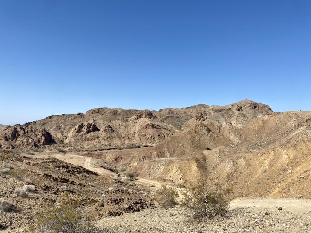 A single-track dirt road winds its way through hilly scrubland. The brown, craggy hills bordering the road sit beneath a clear blue sky.  