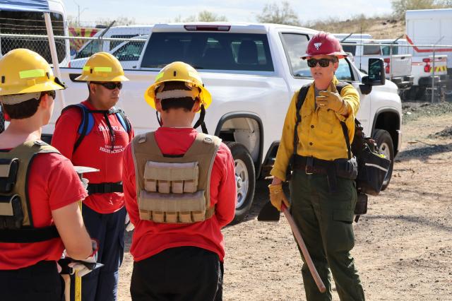 a wildland firefighter in yellow shirt, green pants, and hard hat, speaks to high school students