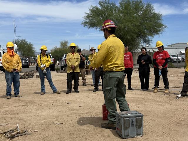 a firefighter in yellow shirt, green pants, and a red hard hat stands in a circle of high school students