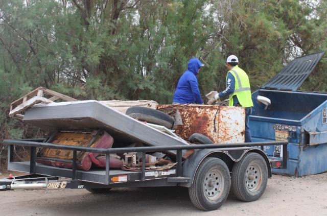 Two men throw refuse from a trailer to into a dumpster.