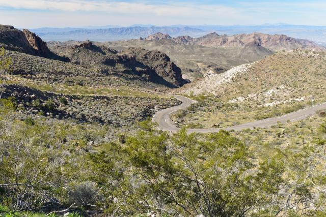 A winding desert road cuts through rugged, shrub-covered mountains, with distant mountain ranges visible under a clear blue sky.