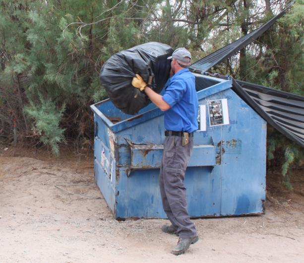 A man throws a bag of trash into a blue dumpster.