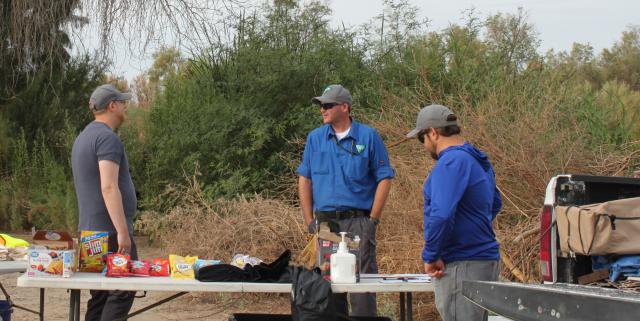 Three men stand around folding tables with snacks and supplies during an outdoor volunteer event near dense desert vegetation.