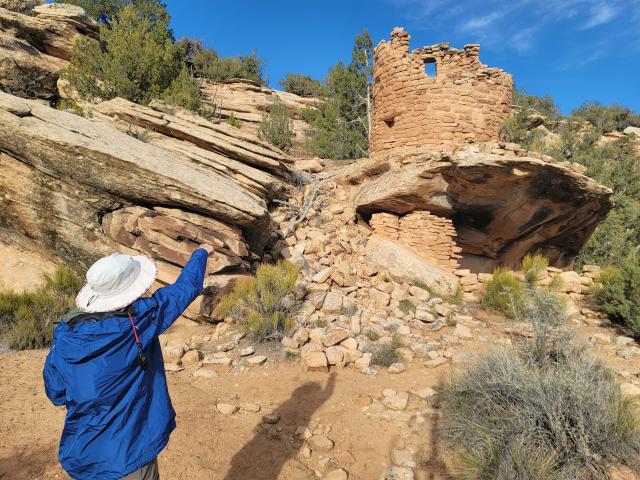 Person in blue jacket and white hat pointing at ancient stone tower built into rocky hillside under clear blue sky.