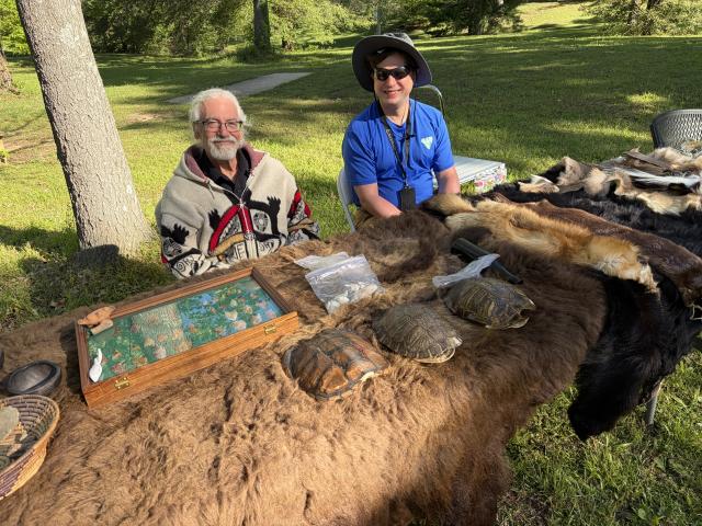 Two people sit at an outdoor table that is covered in animal pelts and archeological artifacts
