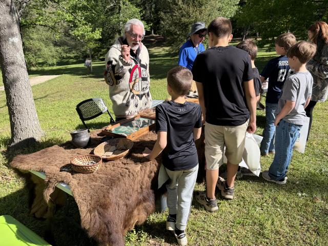 A group of children gathers around BLM table covered with animal pelts and woven baskets, listening to BLM Archaeologist John Sullivan and BLM Planning and Environmental Specialist Joe Edmonds standing behind the display. The table holds artifacts such as pottery pieces and tools.