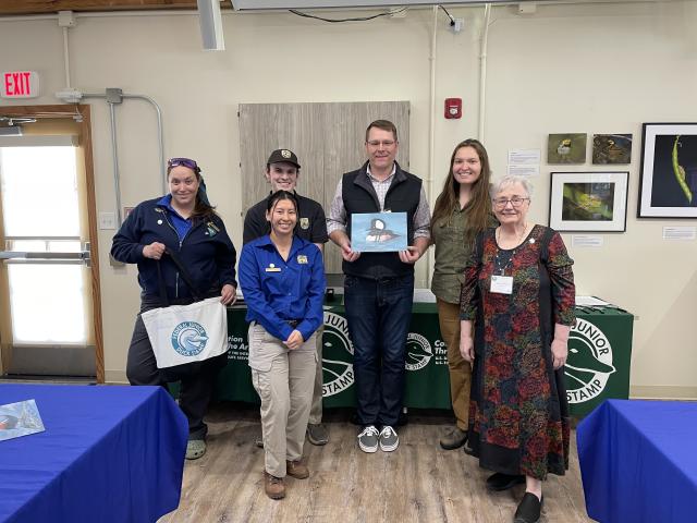 Group of six people standing indoors in front of tables with Federal Junior Duck Stamp banners, one person holding a duck painting.