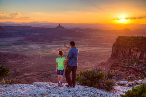 A father and daughter stand on an overlook with landscape features in the distance. 