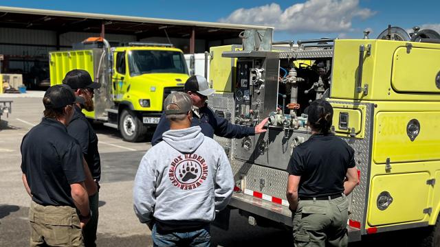 A group of five firefighters, including one wearing an "Elder Rich Weber Cache Box" hoodie, gather around the back of a lime-green fire truck for an equipment demonstration.