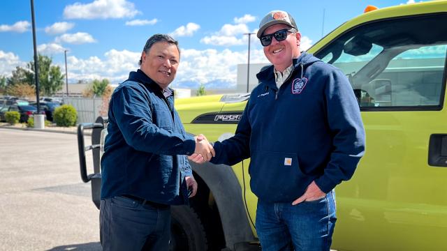 Erik Valdez shakes hands with a smiling Snowville firefighter wearing a navy hoodie and sunglasses in front of a lime-green Ford F-550 wildland engine.