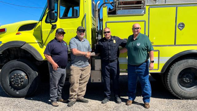 Four men stand together posing for a photo in front of a large lime-green International 7400 fire engine, with two in the center shaking hands.
