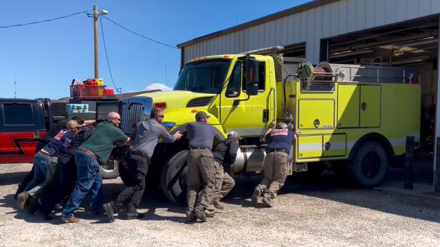 A team of eight people work together to manually push a massive lime-green International wildland fire engine backward into a fire station bay.