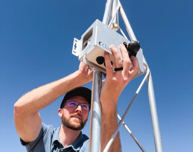 A bearded man in a blue polo adjusts a silver Clarity device on a metal tower under a clear blue sky. He is in a desert setting with a chain-link fence in the background.