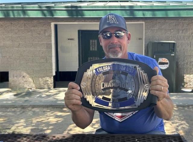 A man wearing sunglasses and a baseball cap holds up a championship-style belt while seated at an outdoor picnic table.