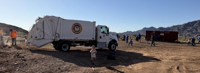 A Yuma County Public Works truck and a large roll-off dumpster sit in a desert cleanup area as volunteers collect trash across rocky public lands near Yuma, Arizona.