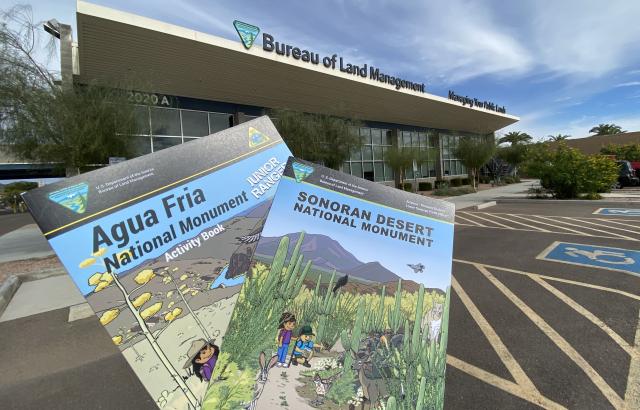 Two illustrated Junior Ranger booklets for Agua Fria and Sonoran Desert National Monuments held in front of the Bureau of Land Management Phoenix District building under a partly cloudy sky.
