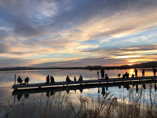 People sitting and standing on a dock at sunrise overlooking calm lake waters with colorful clouds reflecting on the surface.