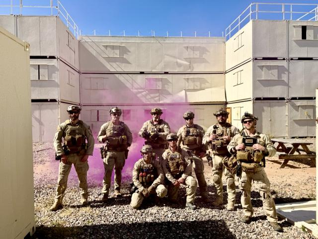 Eight tactical law enforcement officers pose in protective gear outside a training structure as pink marking smoke drifts behind them.