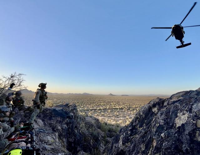 Tactical officers on a rocky ridge signal to an approaching helicopter during a desert rescue training exercise.