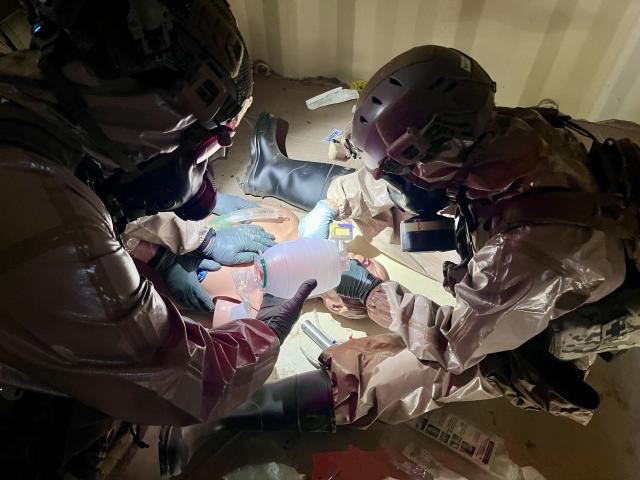 Two tactical officers in protective gear provide simulated emergency care to a training mannequin in a confined space.