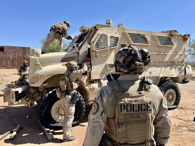 Tactical law enforcement officers train around a tan armored vehicle, with one officer climbing onto the hood.