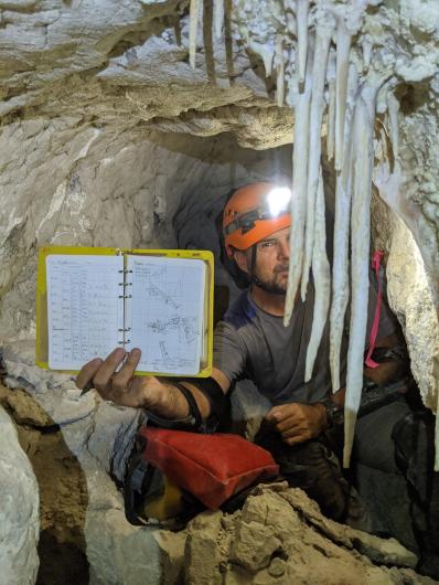 A man in a cave holds up his sketchbook with his notes. 