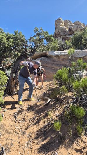Two people using tools to clear a dirt trail on a rocky hillside with shrubs and trees.