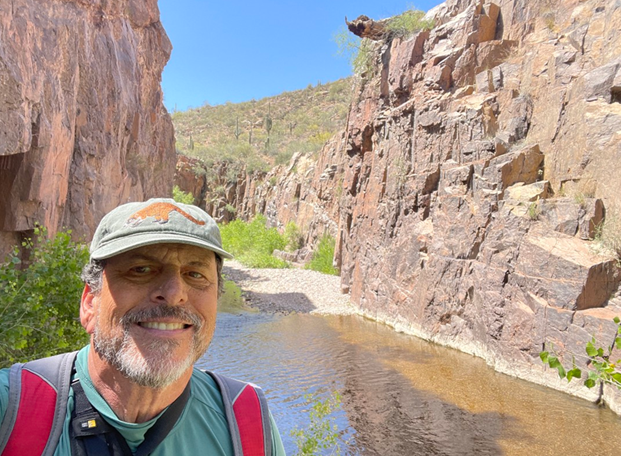 Hiker wearing a hat standing beside a narrow desert canyon stream, with tall rock walls rising on both sides under a bright blue sky.